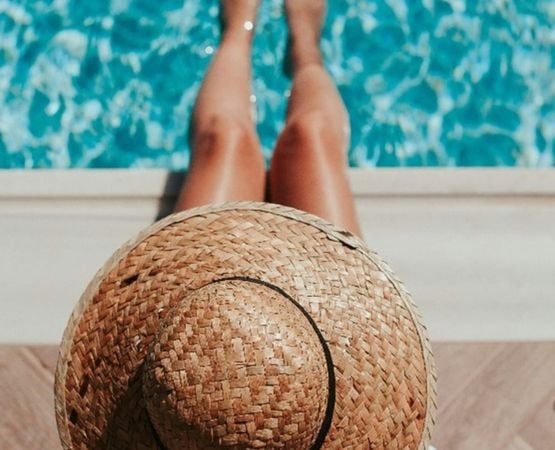Personne assise près d'une piscine, les pieds dans l'eau, portant un chapeau de paille.