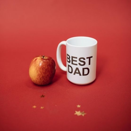 White mug with the inscription "BEST DAD" next to a red apple on a red background.
