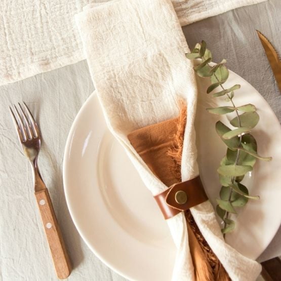 Assiette dressée avec un rond de serviette en cuir tenant une serviette en tissu blanc, accompagnée d'une branche de feuilles vertes et d'une fourchette.