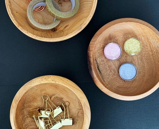 Objets de papeterie et fournitures créatives disposés dans des bols en bois sur fond sombre.