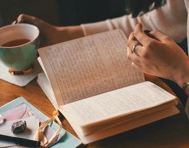 Personne écrivant dans un carnet posé sur une table en bois, accompagnée d’une tasse de thé.