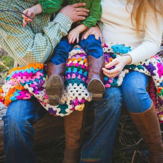 Family seated on a tree trunk, covered in a colourful crocheted blanket; the child is wearing leather boots.