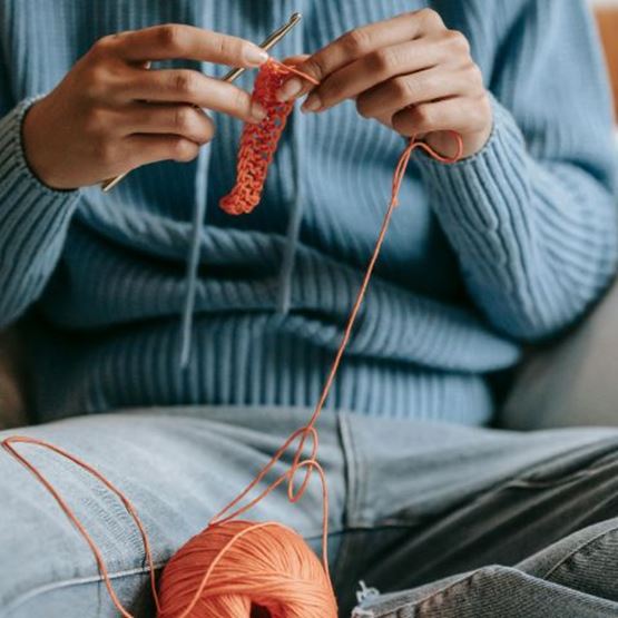 Person in blue jumper sitting, crocheting with orange thread.