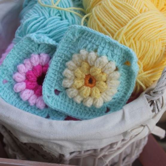 Close-up of a basket containing balls of yellow and blue wool with two crocheted granny squares with floral motifs.