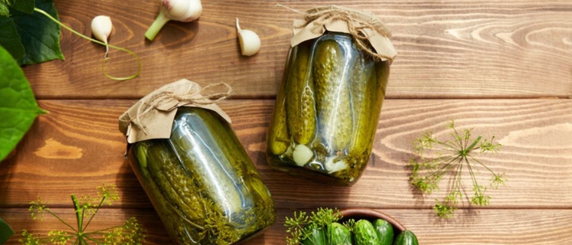 Bocaux de cornichons maison posés sur une table en bois, entourés d’ail et d’herbes.