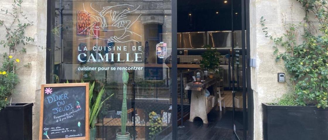 Entrée du lieu « La Cuisine de Camille » avec vitrine, panneau d’ardoise et plantes extérieures.