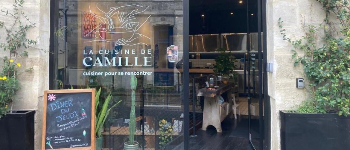 Entrée du lieu « La Cuisine de Camille » avec vitrine, panneau d’ardoise et plantes extérieures.