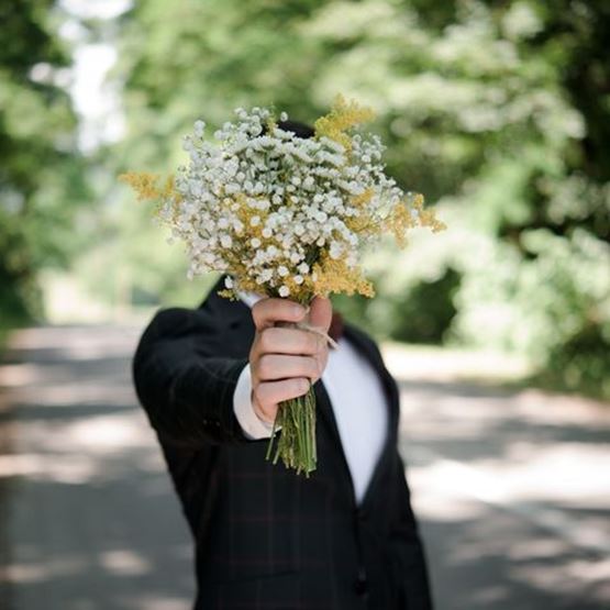 Groom holding a bouquet of gypsophila and yellow flowers in front of him, face hidden.