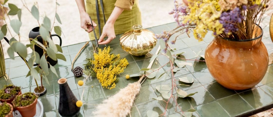 Composition florale en cours de création sur une table avec vases et fleurs séchées.