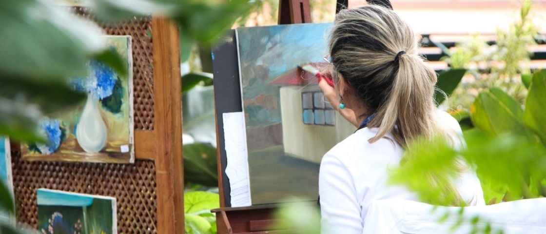 Artiste peignant un tableau en plein air dans un jardin, entourée d’œuvres exposées.