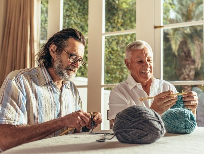 Two senior men knitting together around a brightly lit table, with balls of wool.