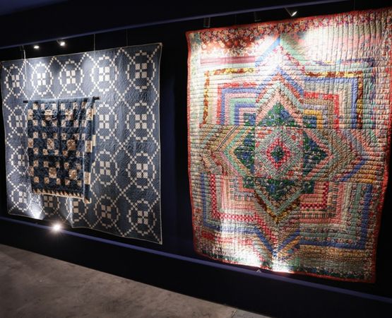 Exposition de quilts colorés et motifs textiles présentés sur un mur dans un espace muséal.
