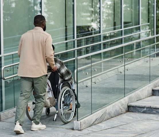 One person helps another to access a building in a wheelchair, using a ramp.