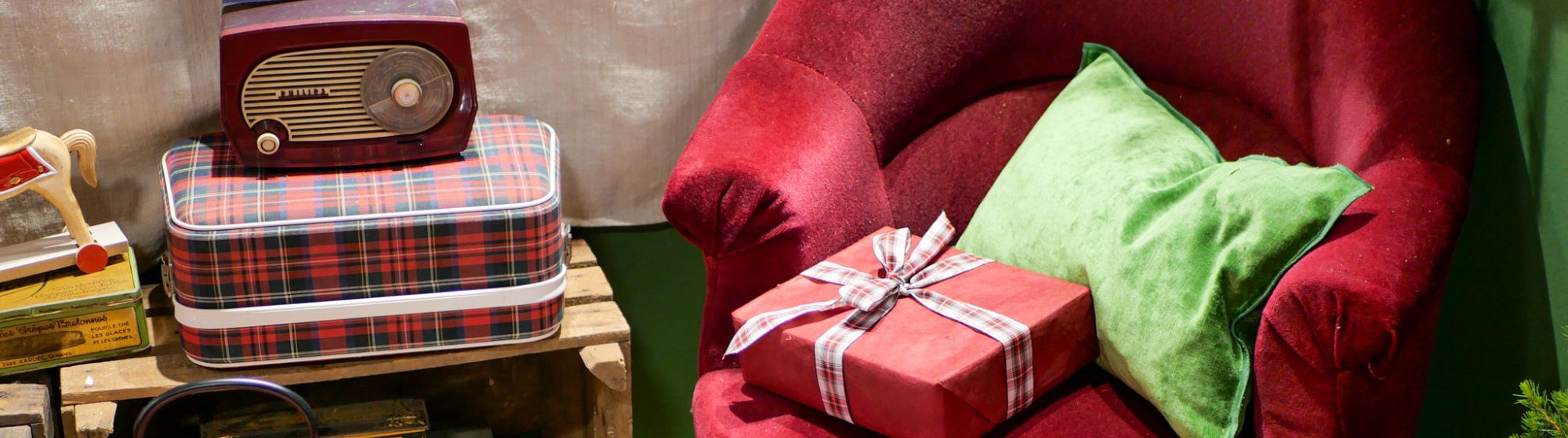 A red Christmas armchair with green cushions, a gift pack, a retro radio, boxes, biscuits and a warm, end-of-year atmosphere.