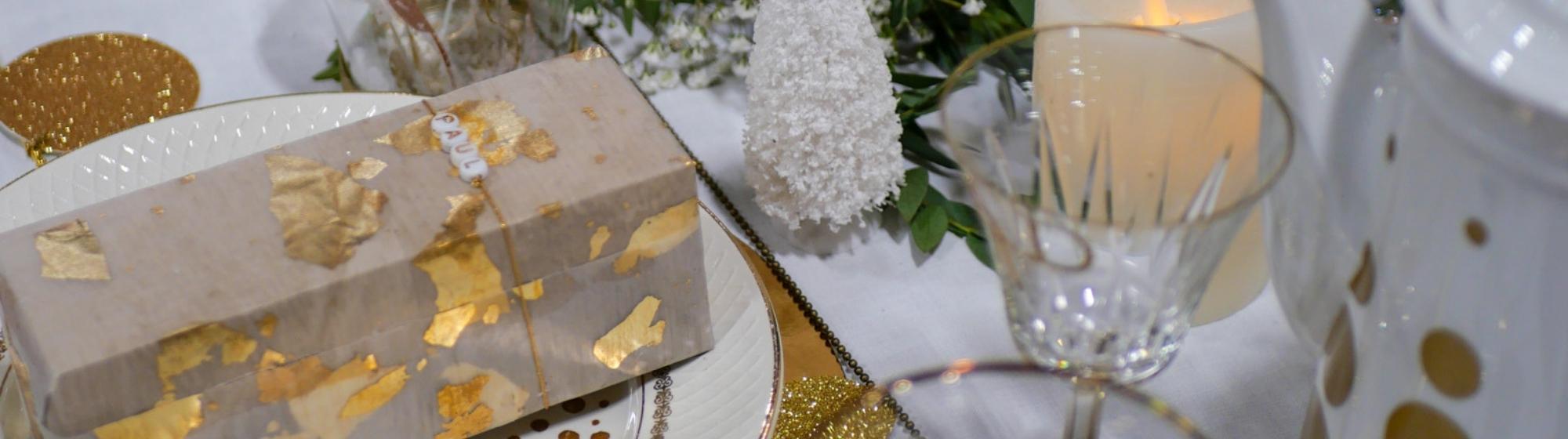 Table de fête décorée de vaisselle blanche et dorée, de verres élégants, de bougies et de feuillage végétal pour une ambiance raffinée.
