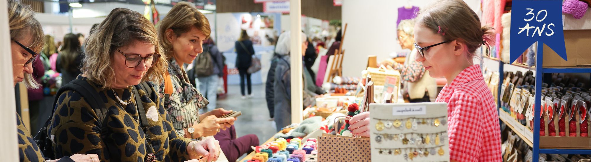 Allée d’un salon créatif avec visiteuses découvrant les stands d’artisanat.