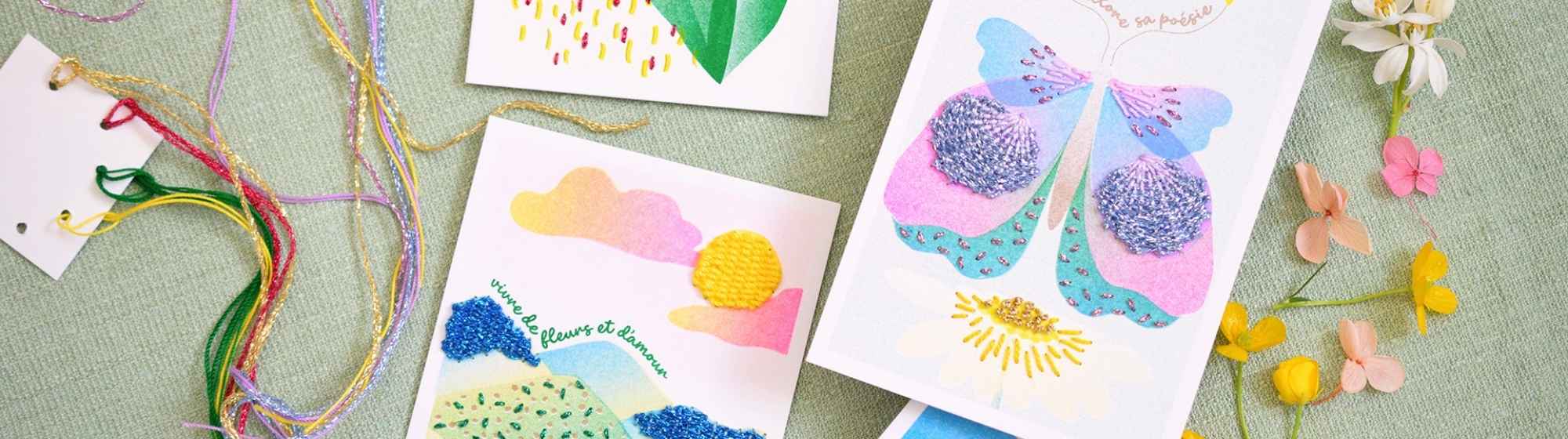 Image format bannière de dessous de verre colorés en laine brodée avec motifs floraux, accompagnés de mugs en céramique bleue et de vases fleuris