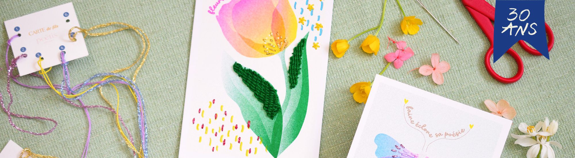 Image format bannière de dessous de verre colorés en laine brodée avec motifs floraux, accompagnés de mugs en céramique bleue et de vases fleuris