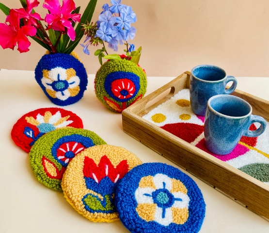Dessous de verre colorés en laine brodée avec motifs floraux, accompagnés de mugs en céramique bleue et de vases fleuris