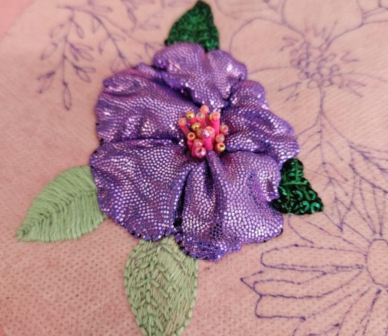 Embroidered violet flower with shiny petals and green leaves on pink fabric, pearl details at centre
