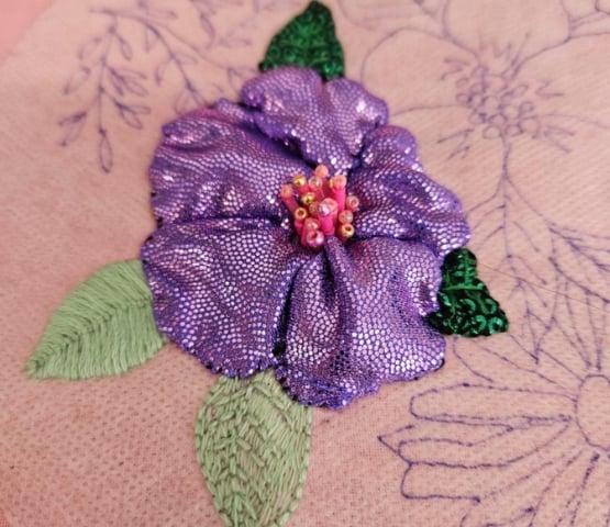 Fleur violette brodée avec pétales brillants et feuilles vertes sur tissu rose, détails en perles au centre