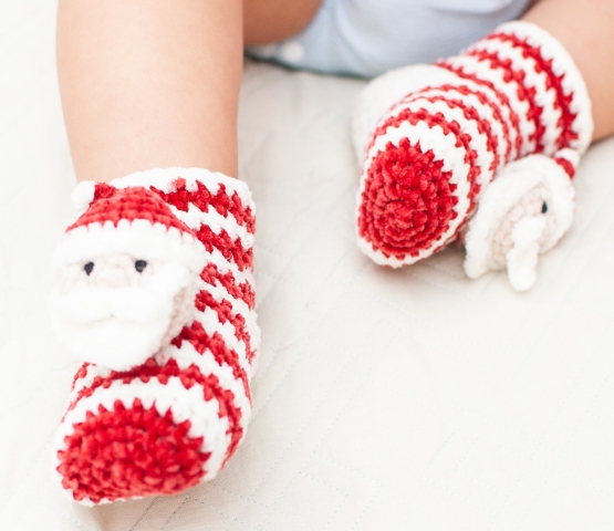 Red and white knitted baby slippers with Santa's head motif, worn by an infant