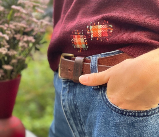 Gros plan sur un pull bordeaux rapiécé avec broderies colorées, porté avec un jean et une ceinture en cuir