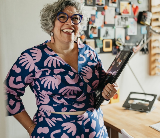 Laura Langlois souriante aux cheveux gris courts, portant un ensemble bleu à motifs roses et des lunettes colorées, tenant un ordinateur portable dans un bureau décoré – format carré