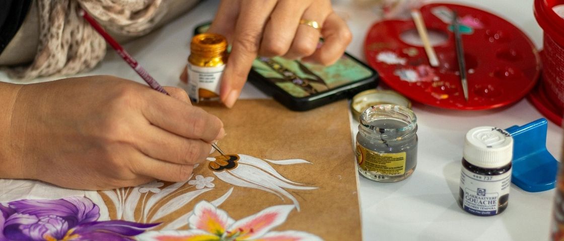 Hand painting coloured flowers with gouache on kraft paper, with pots of paint around them.