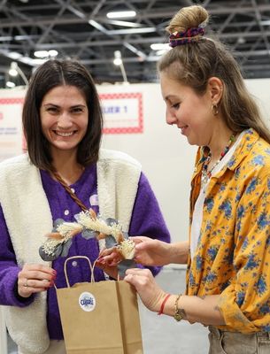 Deux femmes souriantes découvrant des créations artisanales sur un stand de salon créatif.