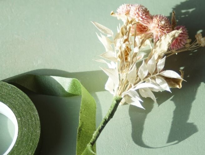 Boutonnière en cours de fabrication avec des fleurs séchées, fixée avec du ruban floral vert.