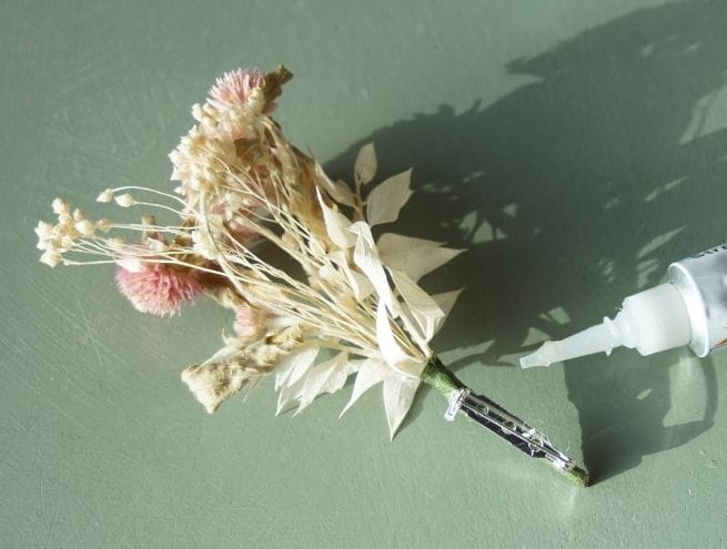 Boutonnière en fleurs séchées terminée avec une attache métallique, à côté d’un tube de colle.