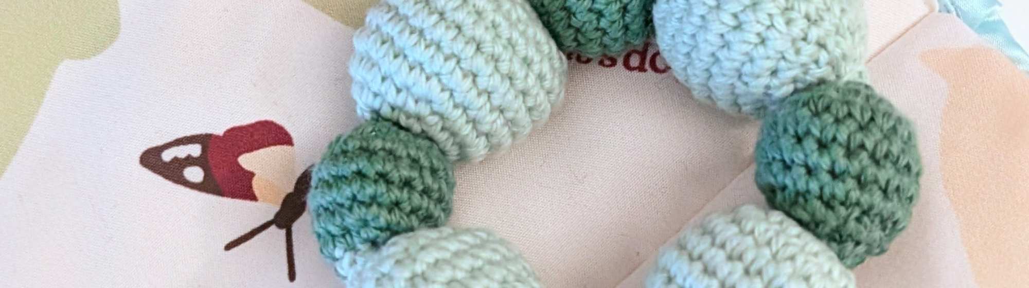 Jouet pour bébé au crochet composé de petites boules vertes et bleu clair assemblées en forme d’anneau, présenté sur un tissu illustré.