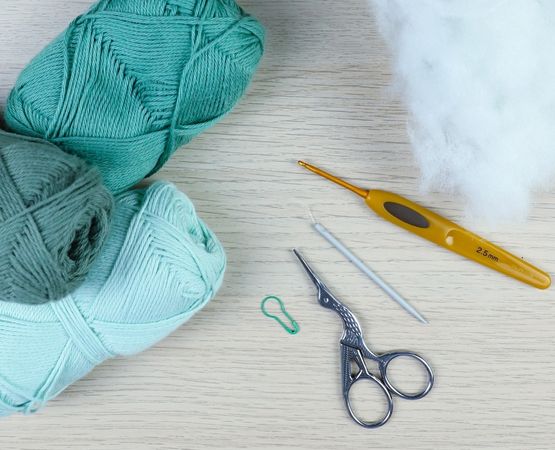 Matériel pour réaliser un jouet bébé au crochet : pelotes de laine vertes, crochet 2,5 mm, ciseaux, aiguille à laine, marqueur de maille et ouate de rembourrage, disposés sur une table en bois clair.