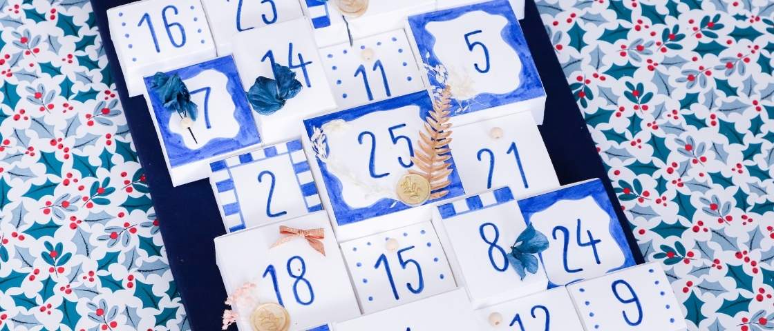 Vue rapprochée d’un calendrier de l’Avent fait main, décoré de motifs bleus, de fleurs séchées et de petits numéros peints.
