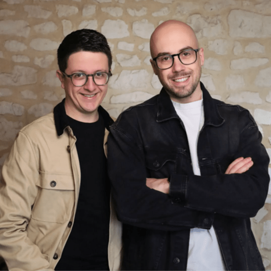 Deux hommes souriants portant des lunettes posent devant un mur en pierre, bras croisés.