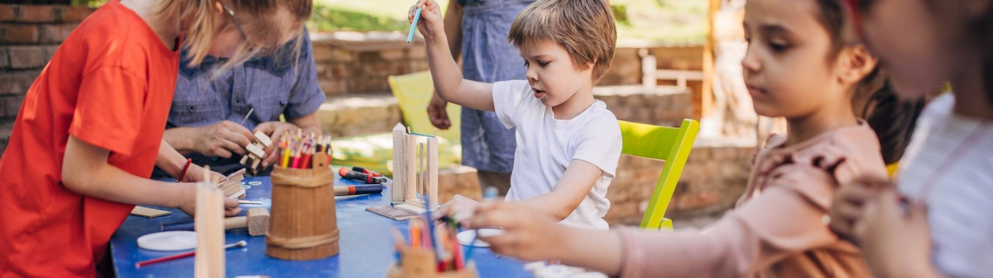 Groupe d’enfants participant à un atelier créatif en plein air, utilisant crayons, bois et outils sur une grande table bleue.