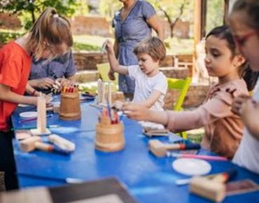 Groupe d’enfants participant à un atelier créatif en plein air, utilisant crayons, bois et outils sur une grande table bleue.