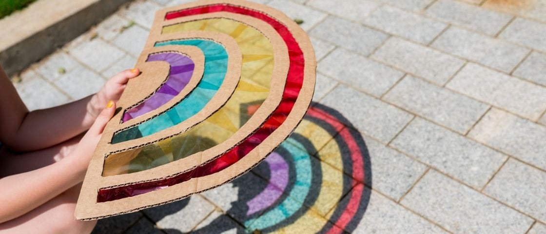 Enfant tenant un attrape-soleil arc-en-ciel en carton coloré, projetant des ombres colorées au sol.