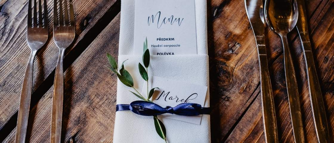 Menu de mariage glissé dans une serviette blanche avec un brin d’olivier, un ruban bleu marine et un marque-place manuscrit.