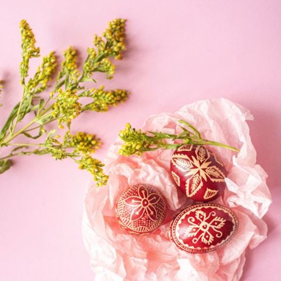 Trois œufs de Pâques rouges richement décorés à la cire, accompagnés de fleurs jaunes sur fond rose.