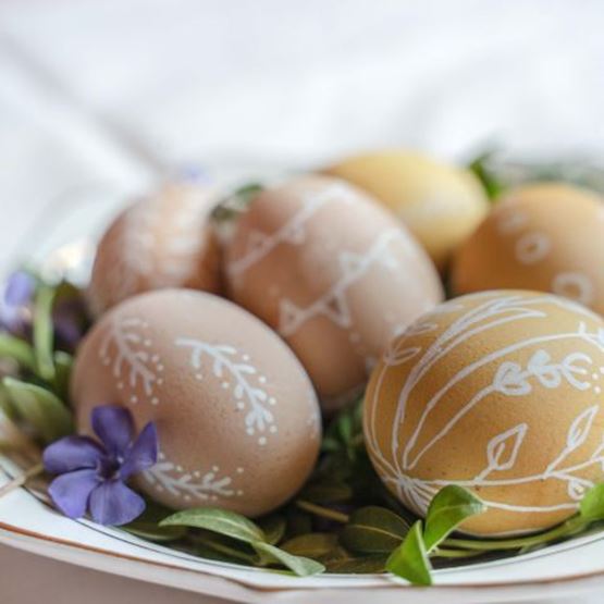 Œufs de Pâques beiges décorés à la main avec des motifs blancs, disposés sur une assiette avec des fleurs violettes.